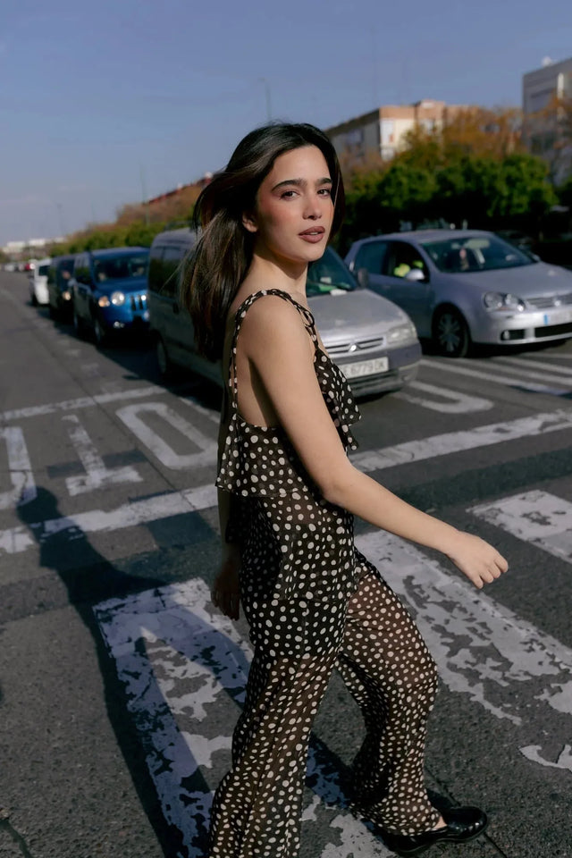Femme marchant dans la rue avec une combinaison fluide à pois blancs et volants, un choix élégant et décontracté pour l'été, accompagnée d'un chemisier voile transparent femme.