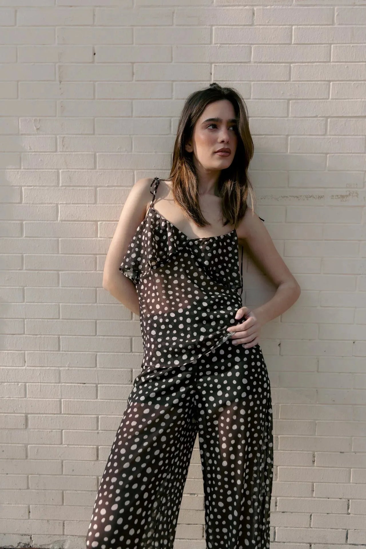 Modèle posant contre un mur avec une combinaison fluide à pois blancs et volants, parfaite pour les journées d'été et à assortir avec un chemisier voile transparent femme.