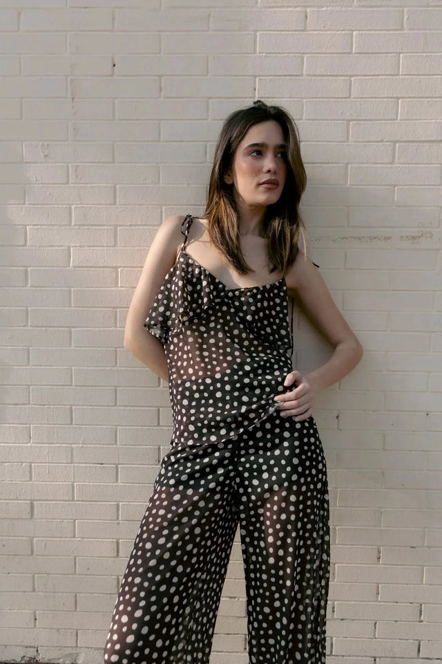 Modèle posant contre un mur avec une combinaison fluide à pois blancs et volants, parfaite pour les journées d'été et à assortir avec un chemisier voile transparent femme.