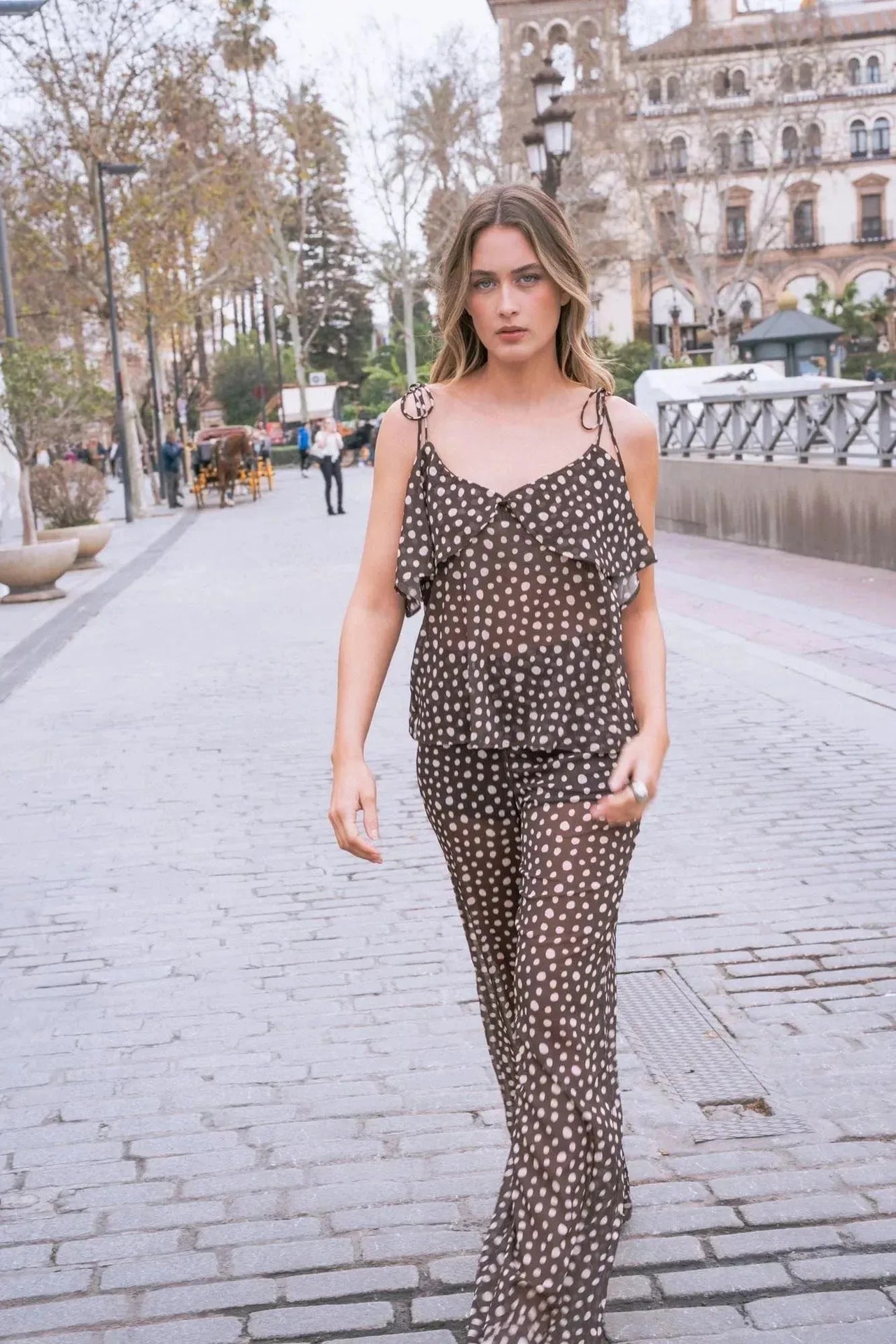 Femme portant une combinaison fluide à pois blancs et volants, marchant sur une promenade, idéale pour un look d'été chic avec un chemisier voile transparent femme.