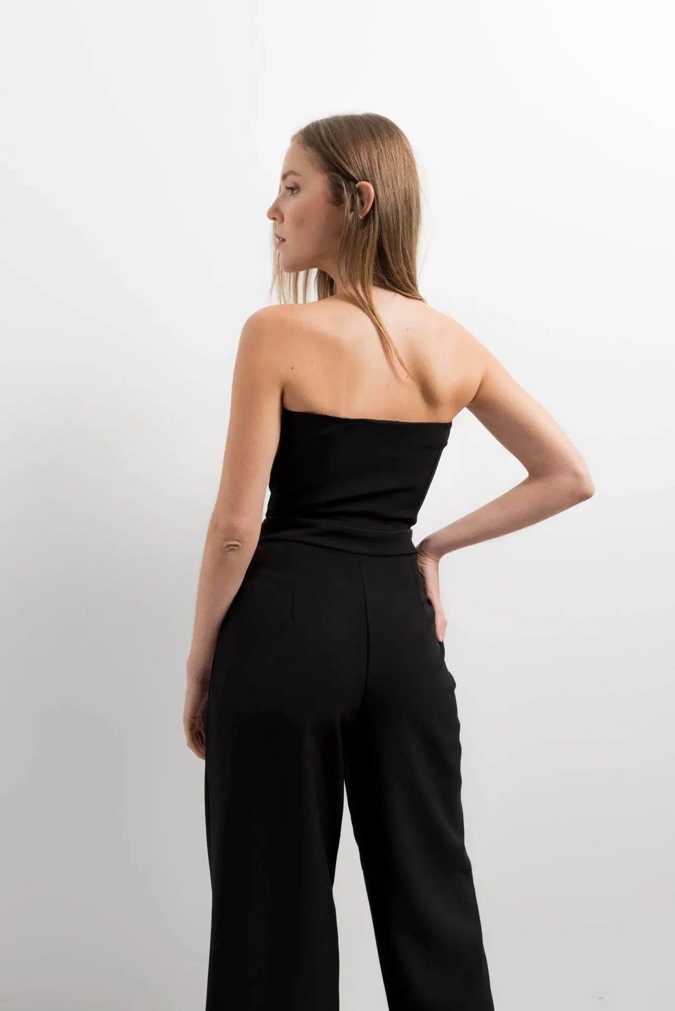 Femme de dos dans une combinaison bustier noir élégante à pantalon fluide, soulignant une coupe moderne et féminine.