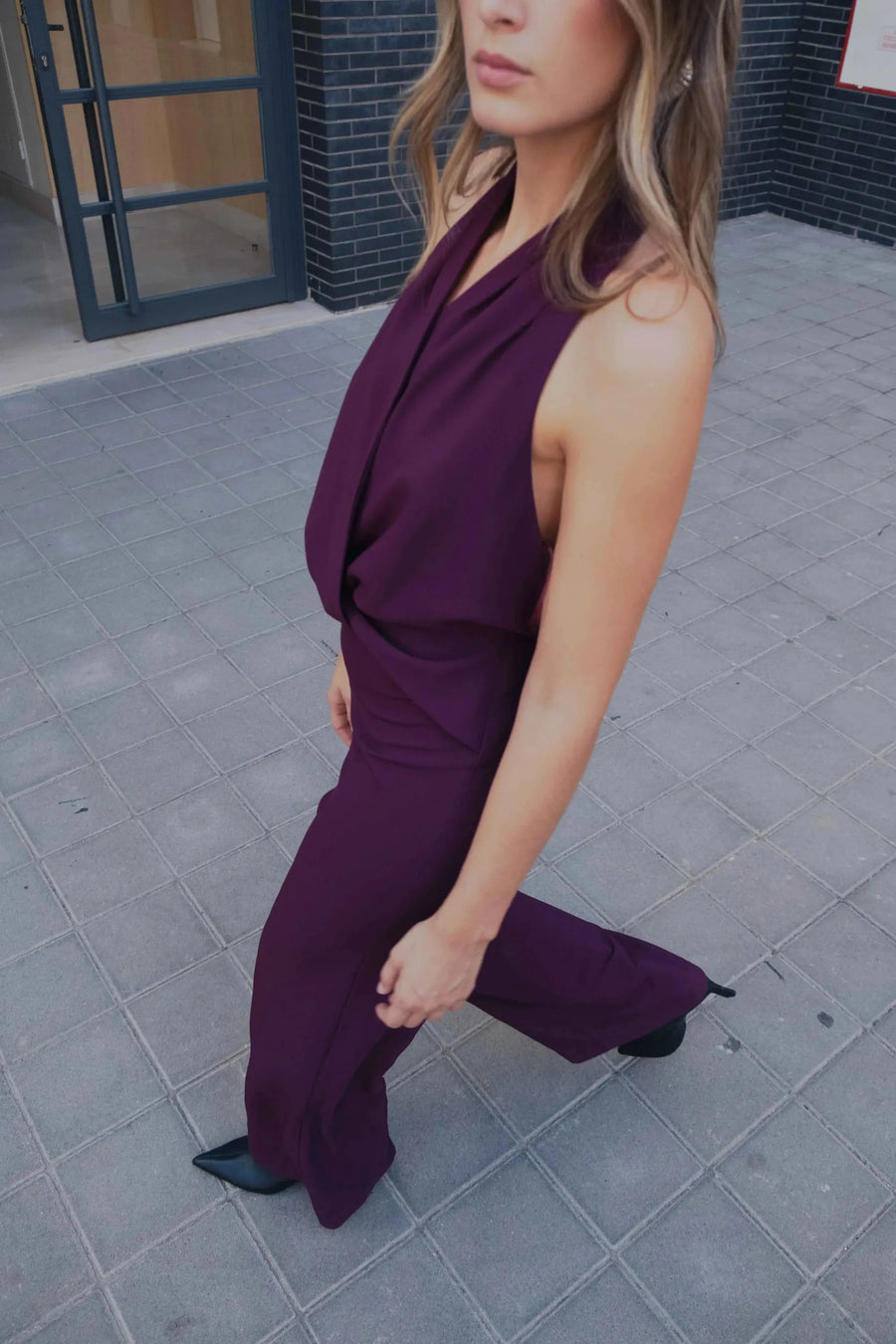 Femme marchant avec assurance dans une combinaison chic pour femme bordeaux prune dos nu à encolure licou sur un sol en pavé.
