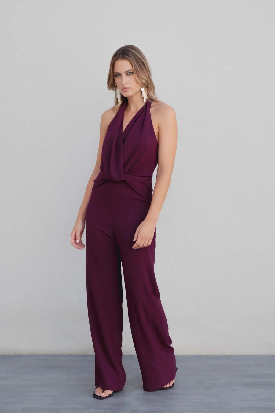 Femme debout en combinaison chic pour femme bordeaux prune dos nu à encolure licou, avec une pose élégante sur fond neutre.
