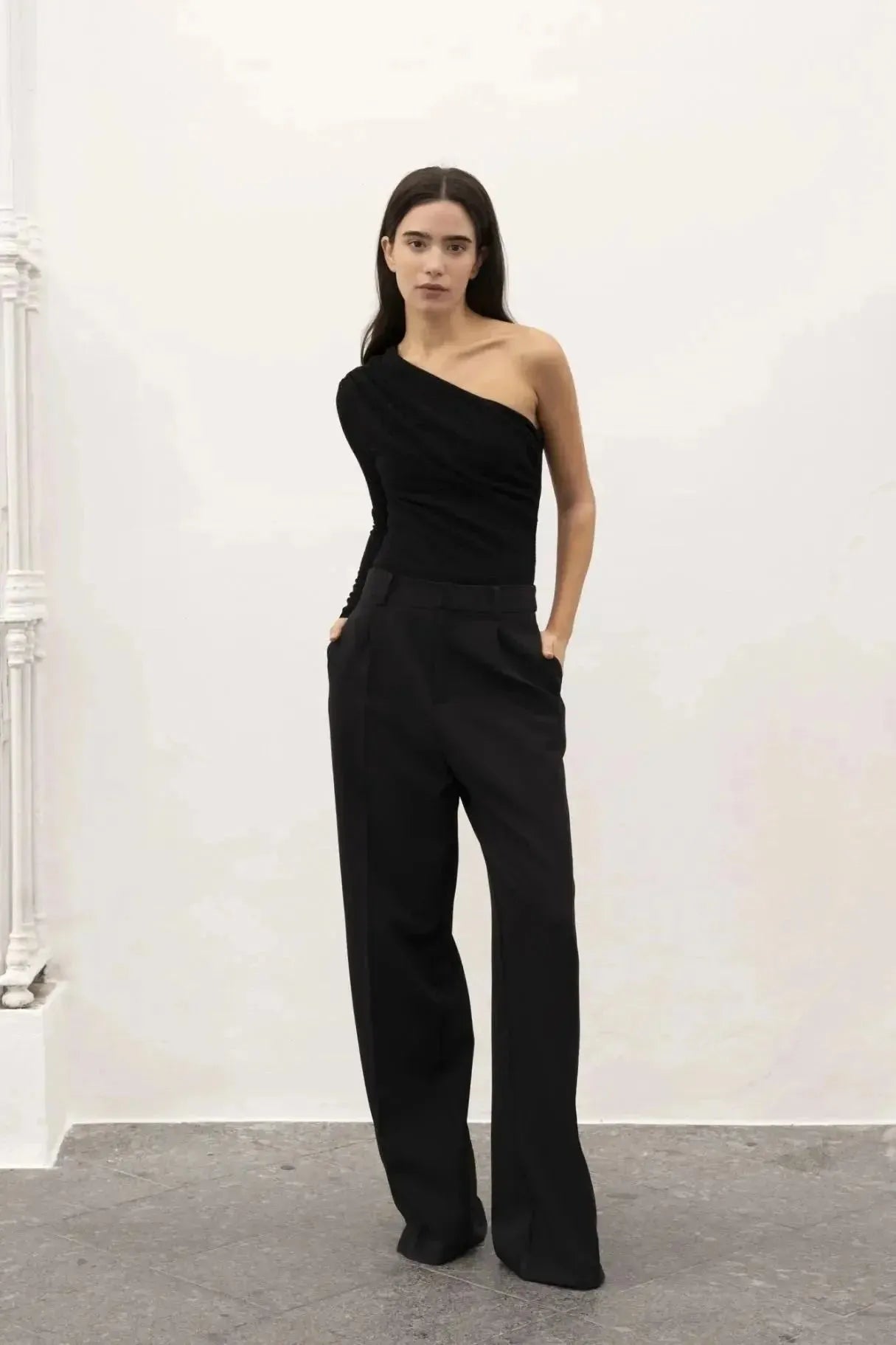 Modèle debout dans un ensemble élégant avec un body asymétrique, portant un pantalon noir ample, dans un environnement minimaliste.