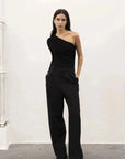 Modèle debout dans un ensemble élégant avec un body asymétrique, portant un pantalon noir ample, dans un environnement minimaliste.