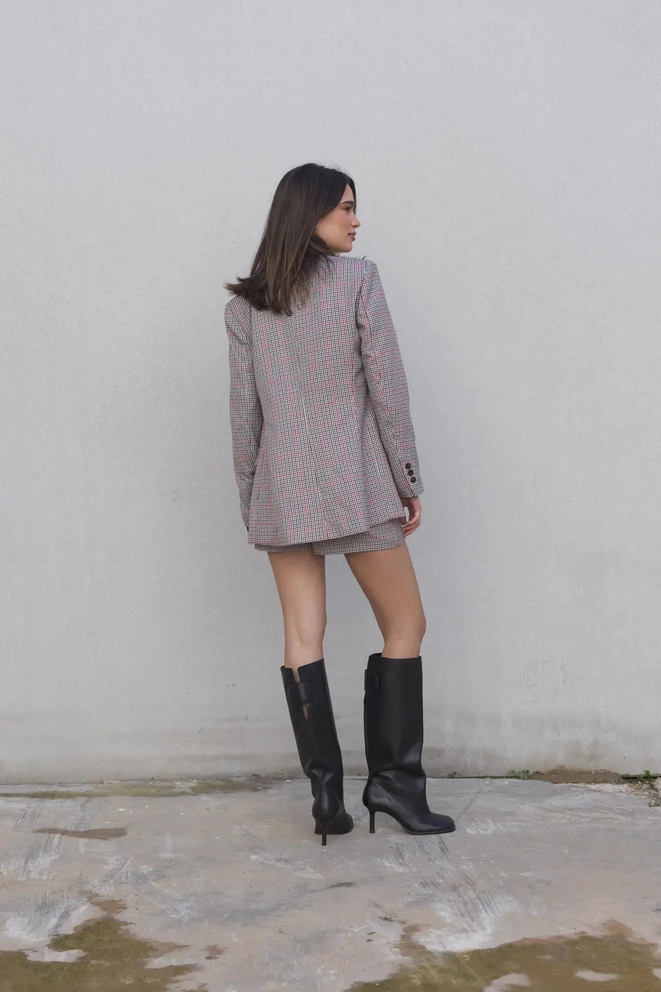 Vue arrière du modèle avec le blazer femme double boutonnage à carreaux et mini jupe élégante de Sapiato, associé à des bottes noires.