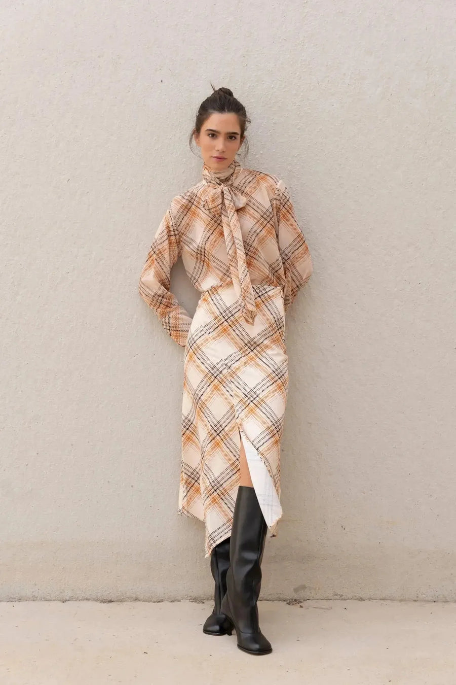 Femme debout avec un chemisier cravate femme à carreaux et des bottes, élégance et confort réunis.