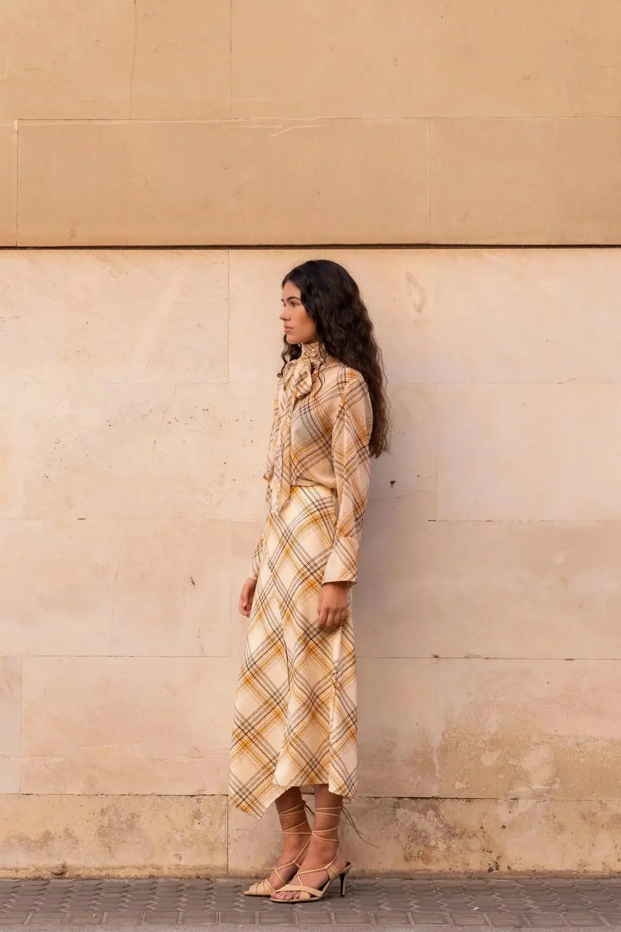 Femme posant côté à un mur avec un chemisier cravate femme à carreaux, soulignant la douceur de la matière.