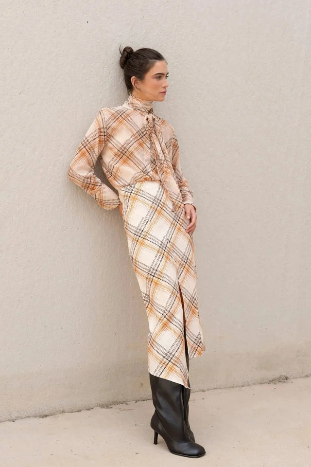 Femme posant contre un mur avec un chemisier cravate femme à carreaux, créant un look chic et moderne.