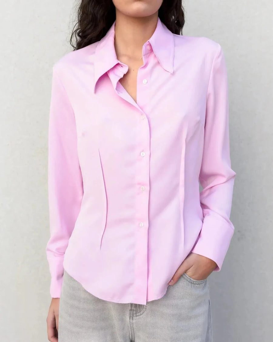 Femme en chemise femme cintrée rose pastel, vêtue de pantalon blanc, marchant dans un décor urbain avec une allure chic.