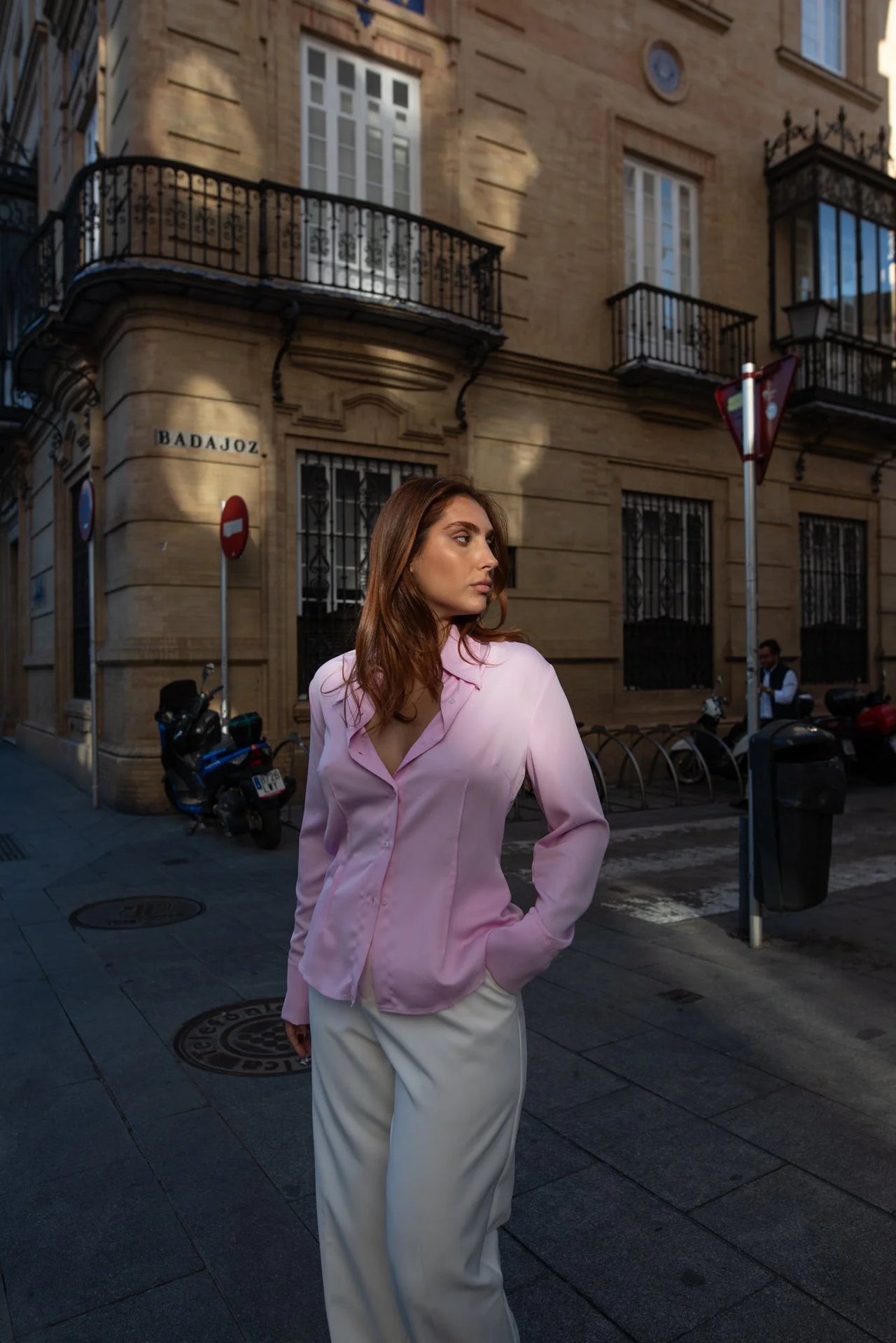 Vue de profil d'une femme en chemise femme cintrée rose pastel, élégante avec pantalon blanc, dans un cadre urbain.