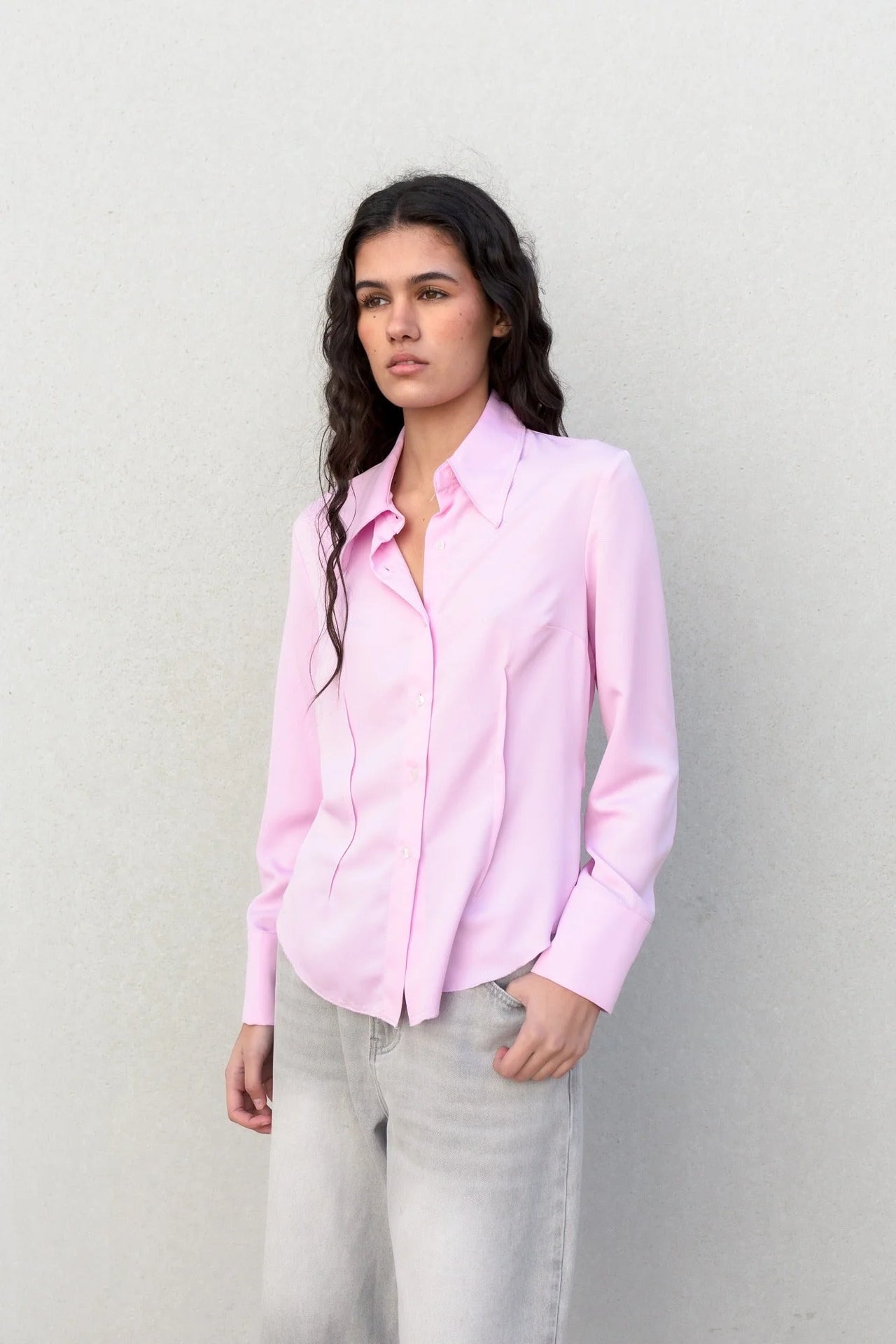 Modèle portant une chemise femme cintrée rose pastel élégante à manches longues avec un style décontracté devant un mur neutre.