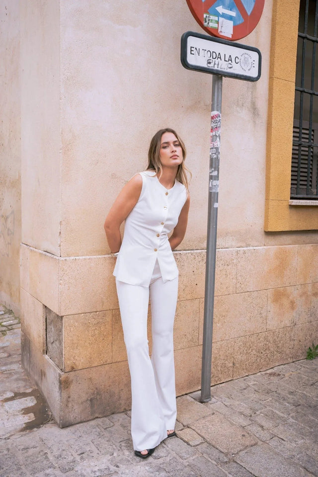 Femme avec une blouse chic femme sans manches à boutons dorés, stylée, posant contre un mur, idéale pour des occasions professionnelles et casual.
