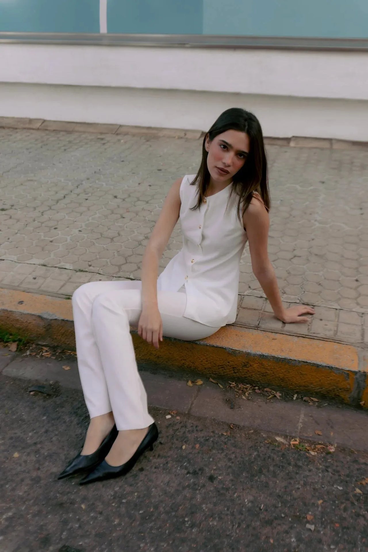 Femme assise sur le bord d'un trottoir, avec une blouse chic femme sans manches à boutons dorés, ajoutant une touche de sophistication à son style décontracté.