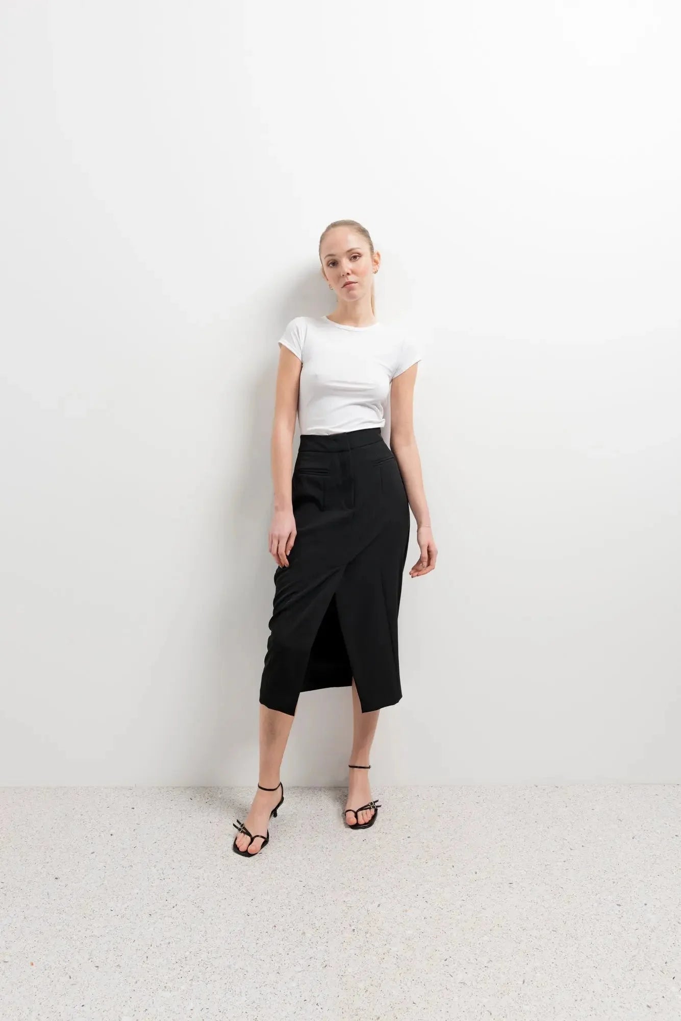 Femme stylée en jupe noire fendue, combinée avec un t-shirt blanc et des sandales pour un look décontracté.