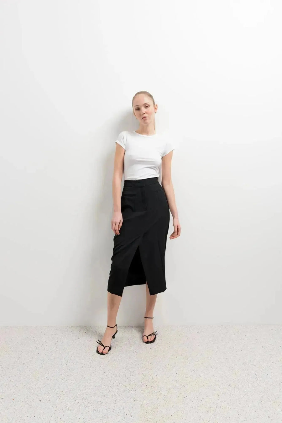 Femme stylée en jupe noire fendue, combinée avec un t-shirt blanc et des sandales pour un look décontracté.