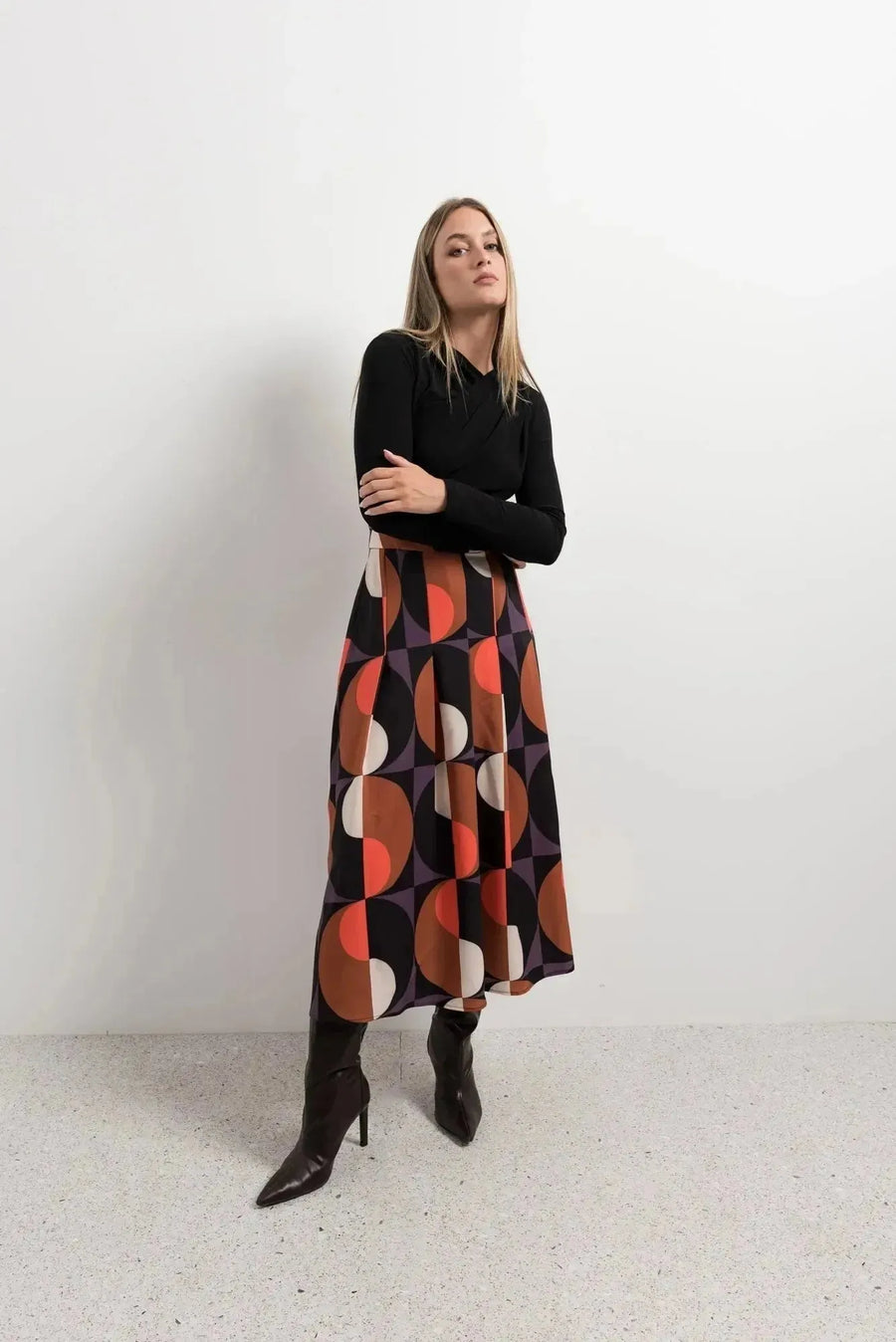 Femme portant une jupe plissée imprimée géométrique, stylée avec un haut noir. Cette jupe année 70 apporte une touche rétro et moderne à son look.