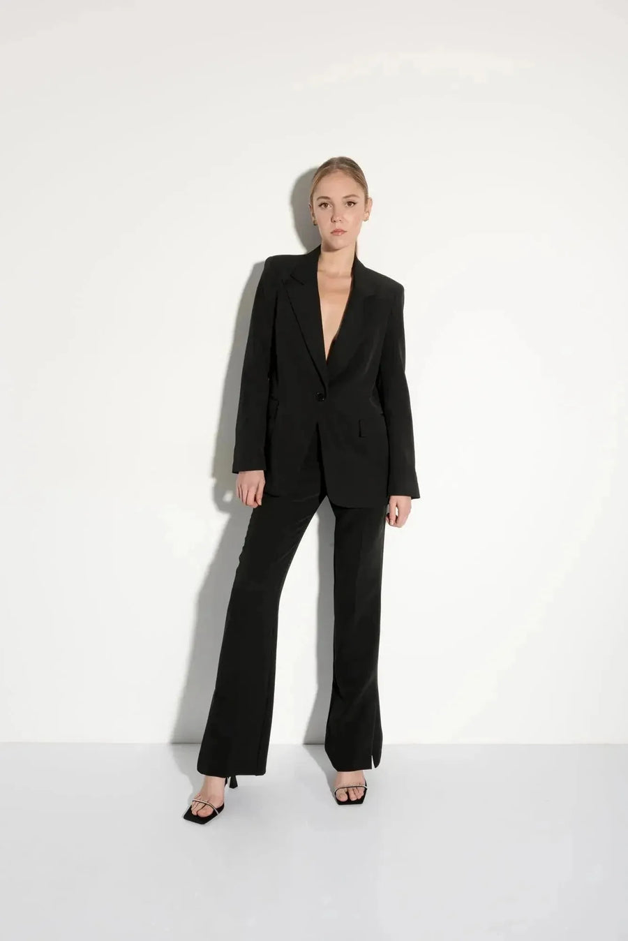 Pantalon Satin Noir à Ourlets Fendus – Confort Élégant avec Poches et Taille Élastique, mannequin posant en blazer noir, une option chic pour un pantalon féminin polyvalent.