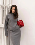 Femme en jupe maille grise, confortable et élégante, en position décontractée avec un pull gris et un sac rouge, le regard posé sur l'objectif.