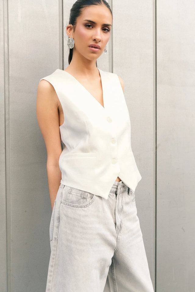 Femme portant un gilet blanc sans manches chic coupe tailleur, avec des boutons ton sur ton, associé à un jean délavé.