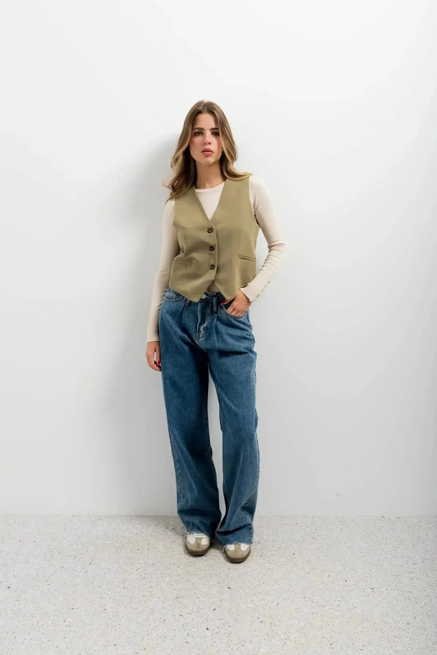 Femme portant le gilet élégant vert facile à assortir, un gilet court sans manche femme, avec un top beige, des jeans amples et des chaussures argentées.