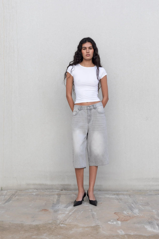 Femme posant avec style en bermuda jean femme gris clair, oversize et à taille haute, associé à un t-shirt blanc, parfait pour un look décontracté.