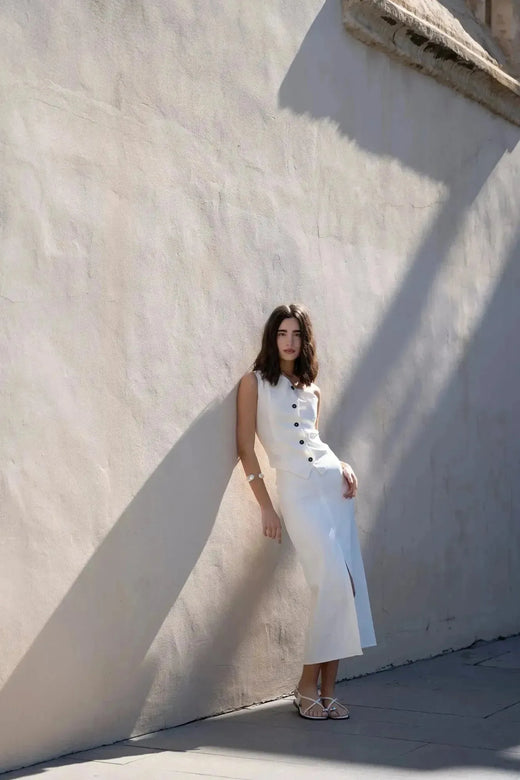 Femme stylée portant une jupe blanche longue femme fendue arrière, posant contre un mur, mettant en avant une élégante coupe flatteuse.