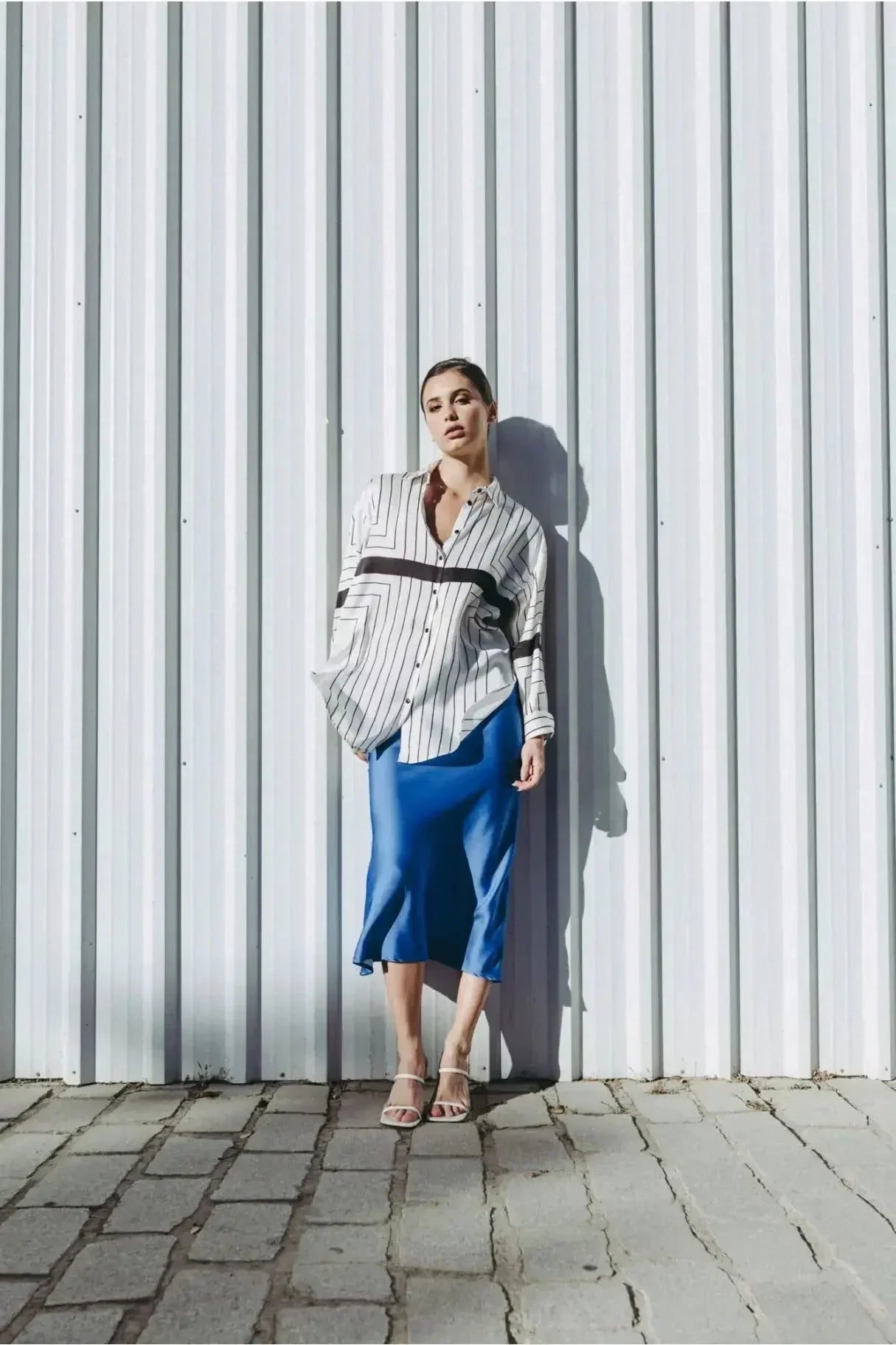Femme stylée affichant une jupe bleue électrique midi satinée avec une chemise rayée, parfaite pour une allure moderne et élégante.