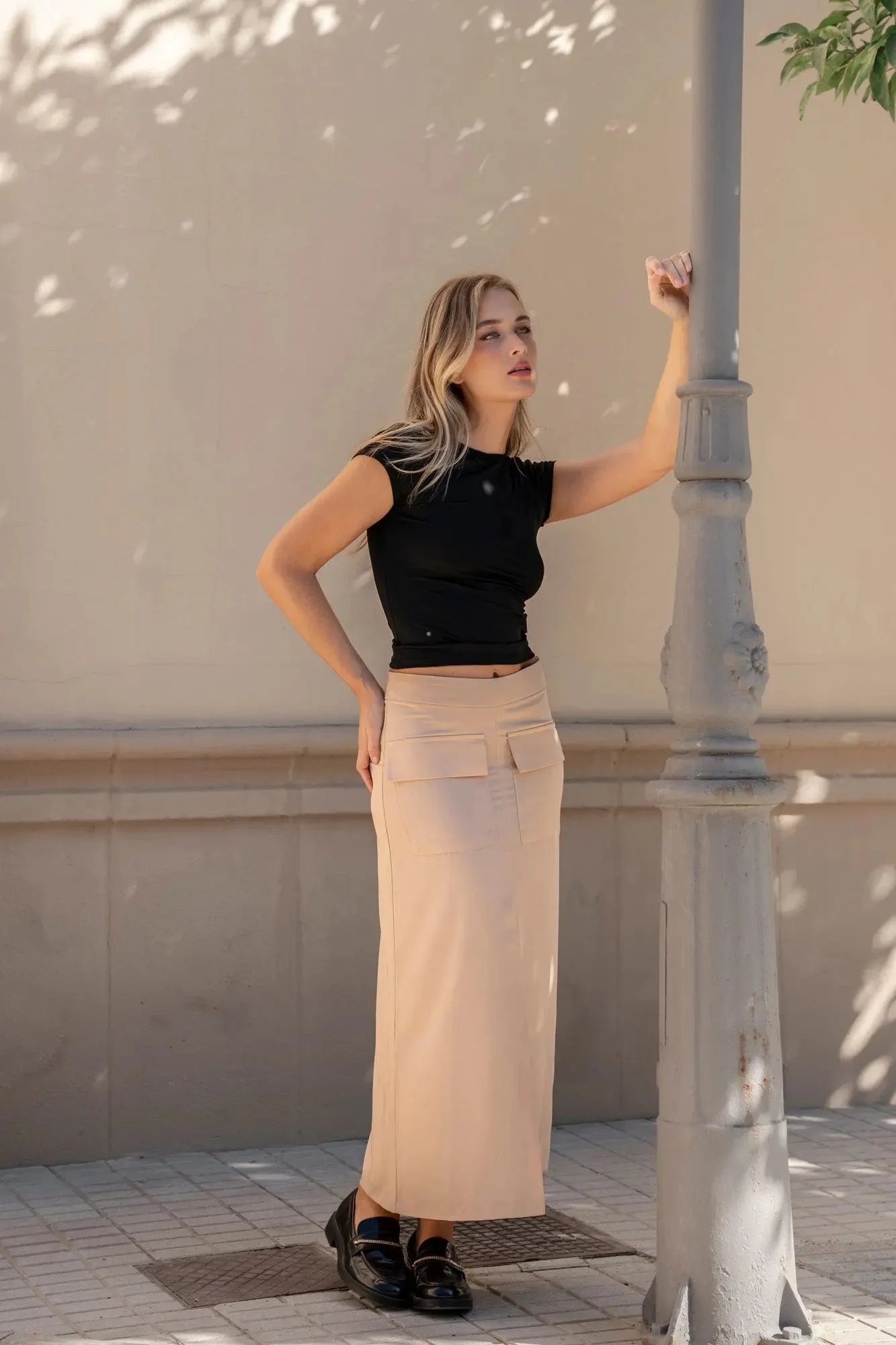 Femme élégante portant la jupe cargo femme beige, avec un haut noir, posant contre un lampadaire en extérieur.