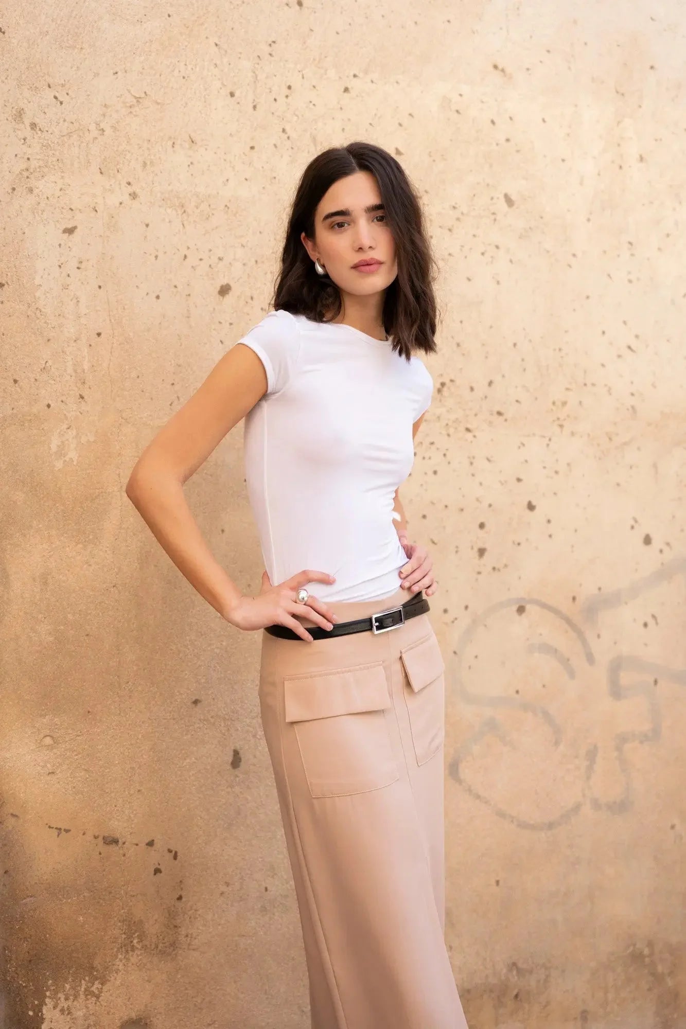Femme posant en jupe cargo femme beige avec un haut blanc, avec un mur texturé en arrière-plan.