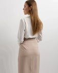 Vue arrière d'une femme en jupe longue beige fendue chic élastique taille haute, montrant un look raffiné avec une chemise blanche.