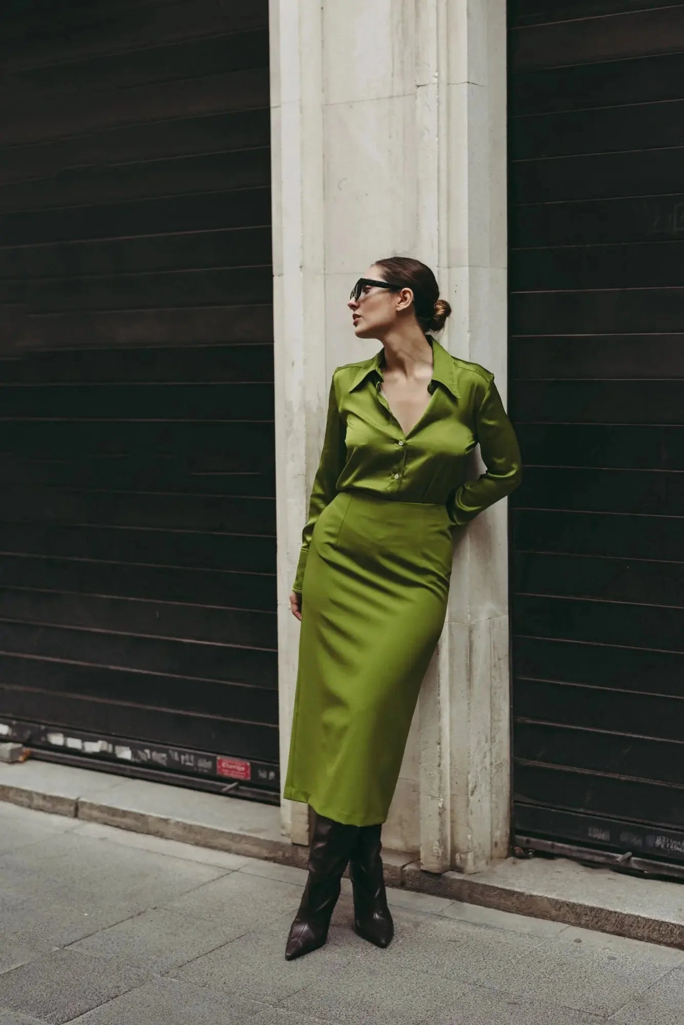 Femme élégante dans une jupe verte longue avec fente latérale, se reposant contre un mur, montrant un look moderne et tendance.