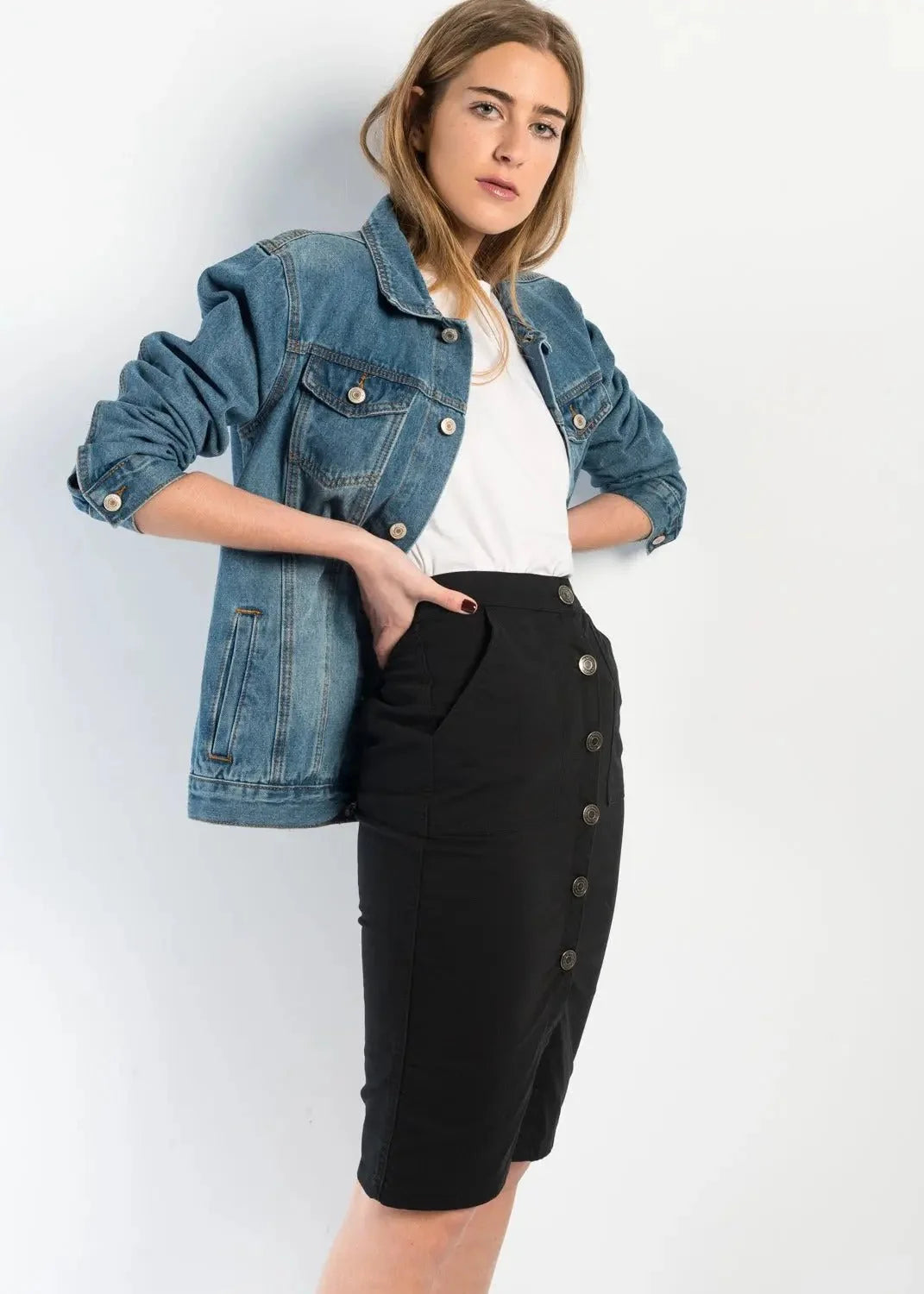 Femme avec une jupe bouton devant noire en denim, posant avec une veste en jean, soulignant l'attrait polyvalent et durable de la jupe.