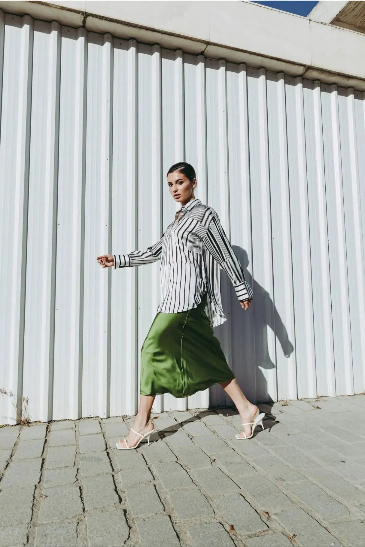 Femme marchant avec confiance en jupe verte satin et chemise à rayures, montrant l'élégance du style.