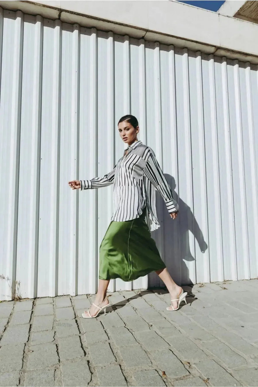 Femme marchant avec confiance en jupe verte satin et chemise à rayures, montrant l'élégance du style.