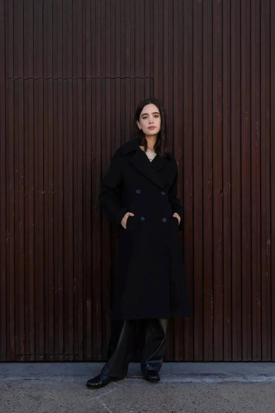 Un manteau long chaud femme noir à double boutonnage porté par une femme, avec un look chic et moderne, devant un mur en bois.