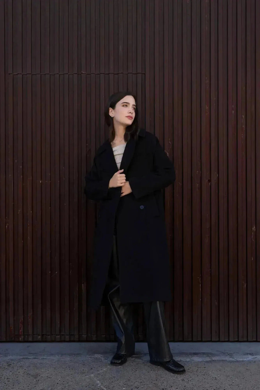 Femme dans un manteau long chaud femme noir à double boutonnage, en train de poser avec une allure décontractée devant un fond en bois.