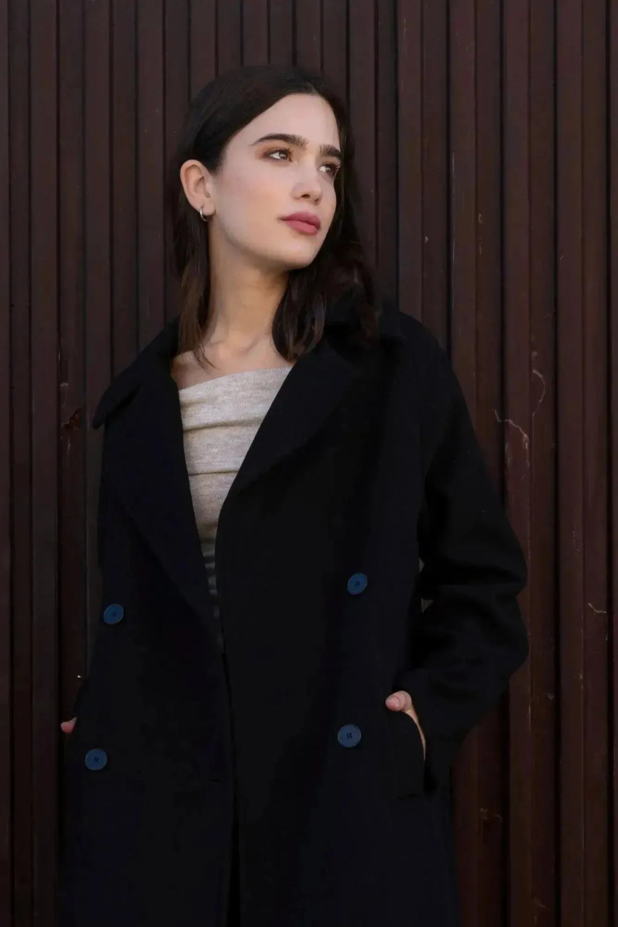 Femme stylée dans un manteau long chaud femme noir à double boutonnage, posant avec une allure chic contre un mur en bois.