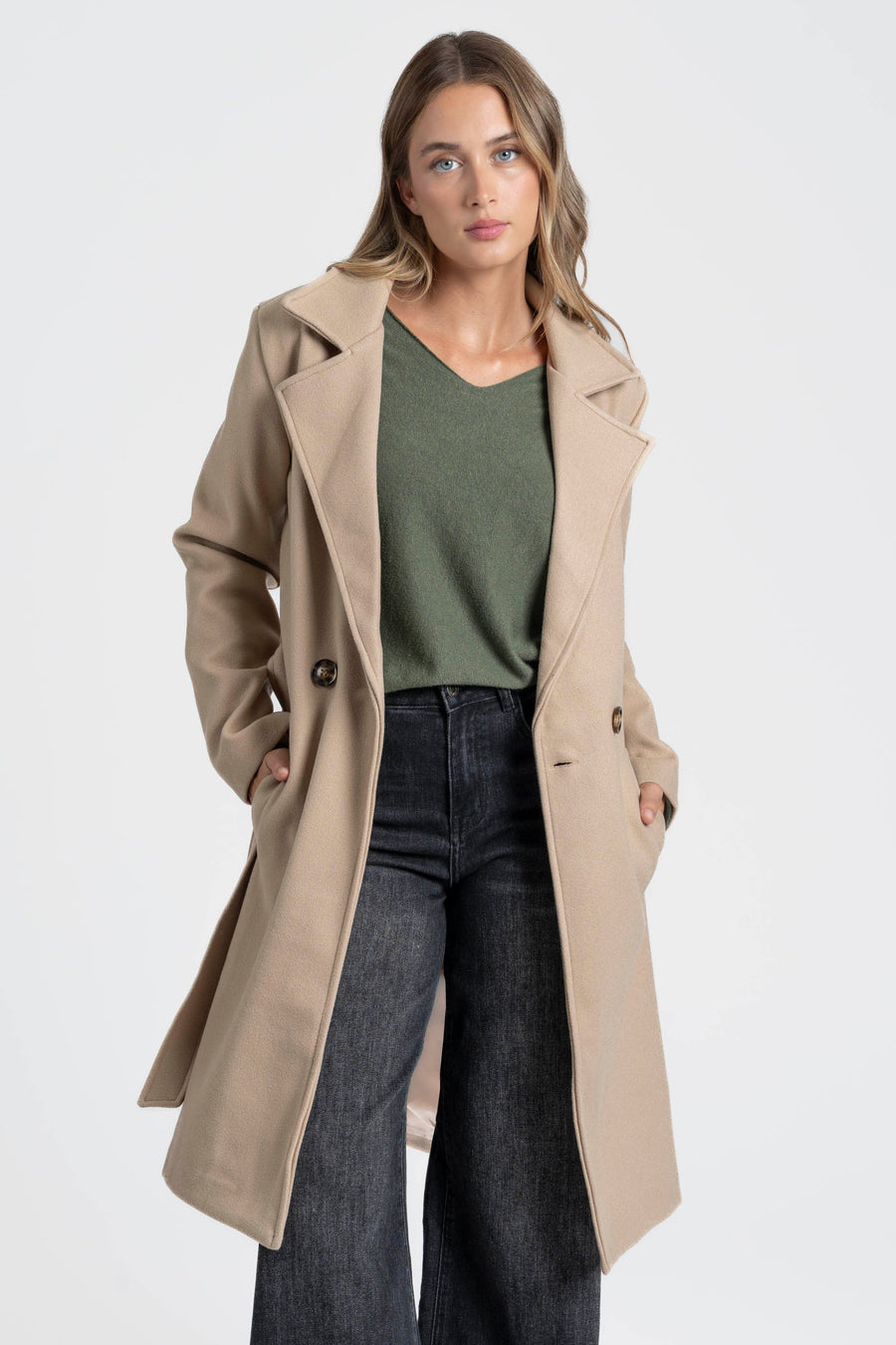 Manteau beige porté ouvert sur un haut vert, mains dans les poches