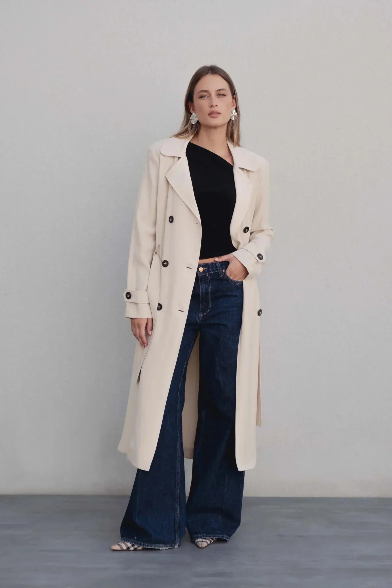 Femme portant un manteau trench femme fluide long beige, associé à un top noir et un jeans évasé. Élégance décontractée avec ceinture.