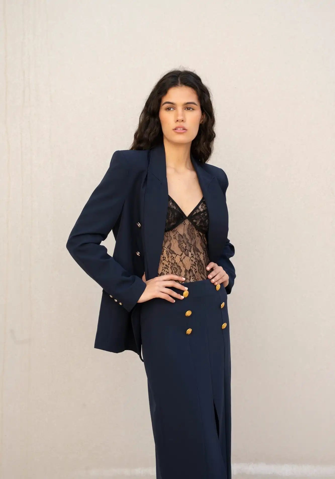 Blazer double-breasted bleu marine avec une jupe longue flashy assortie, ornée de boutons en or et d'une fente frontale, porté avec un body en dentelle noire.