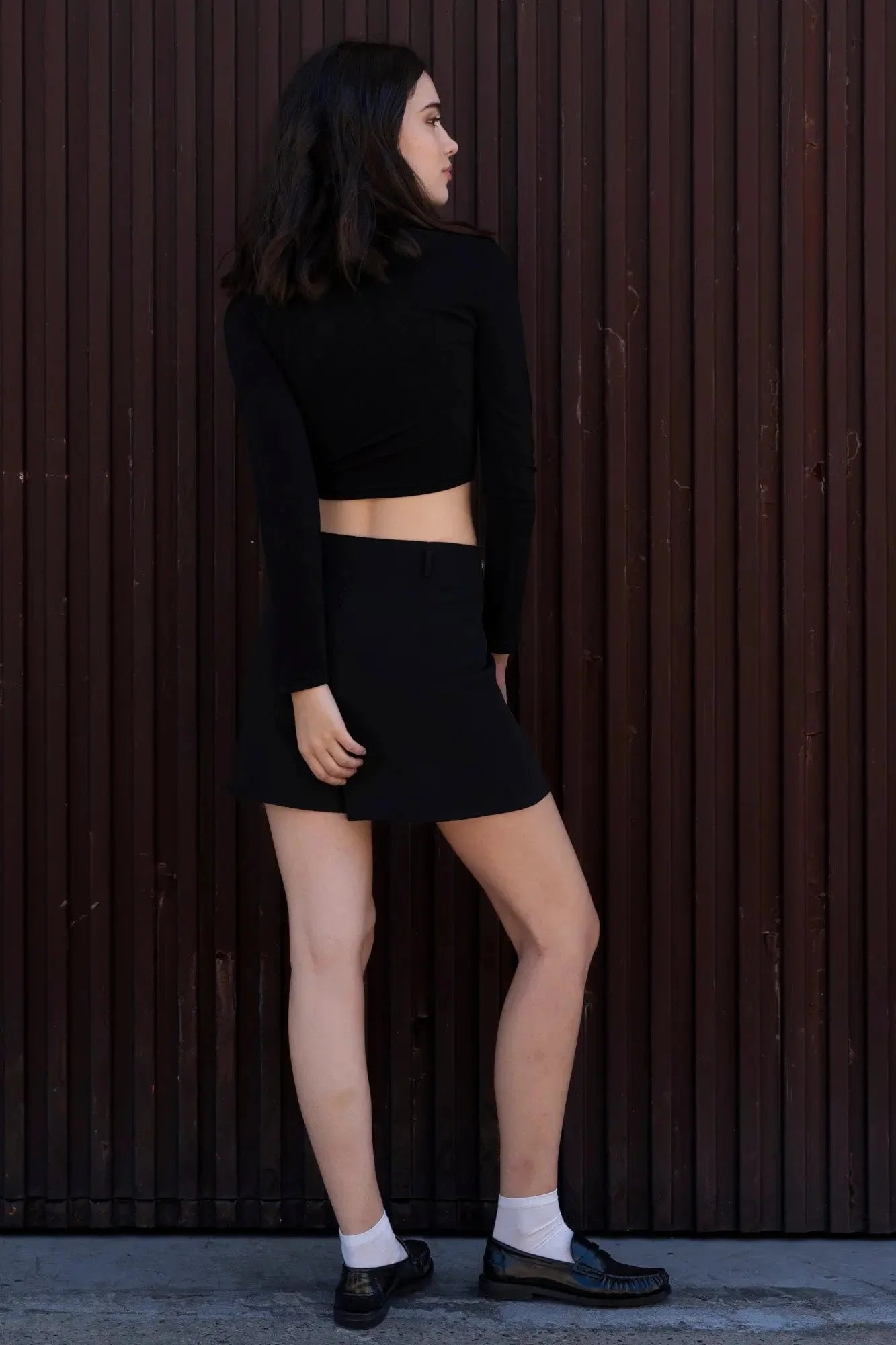 Femme vue de profil, portant une mini jupe noir fendue avec un haut noir, devant un mur en bois.