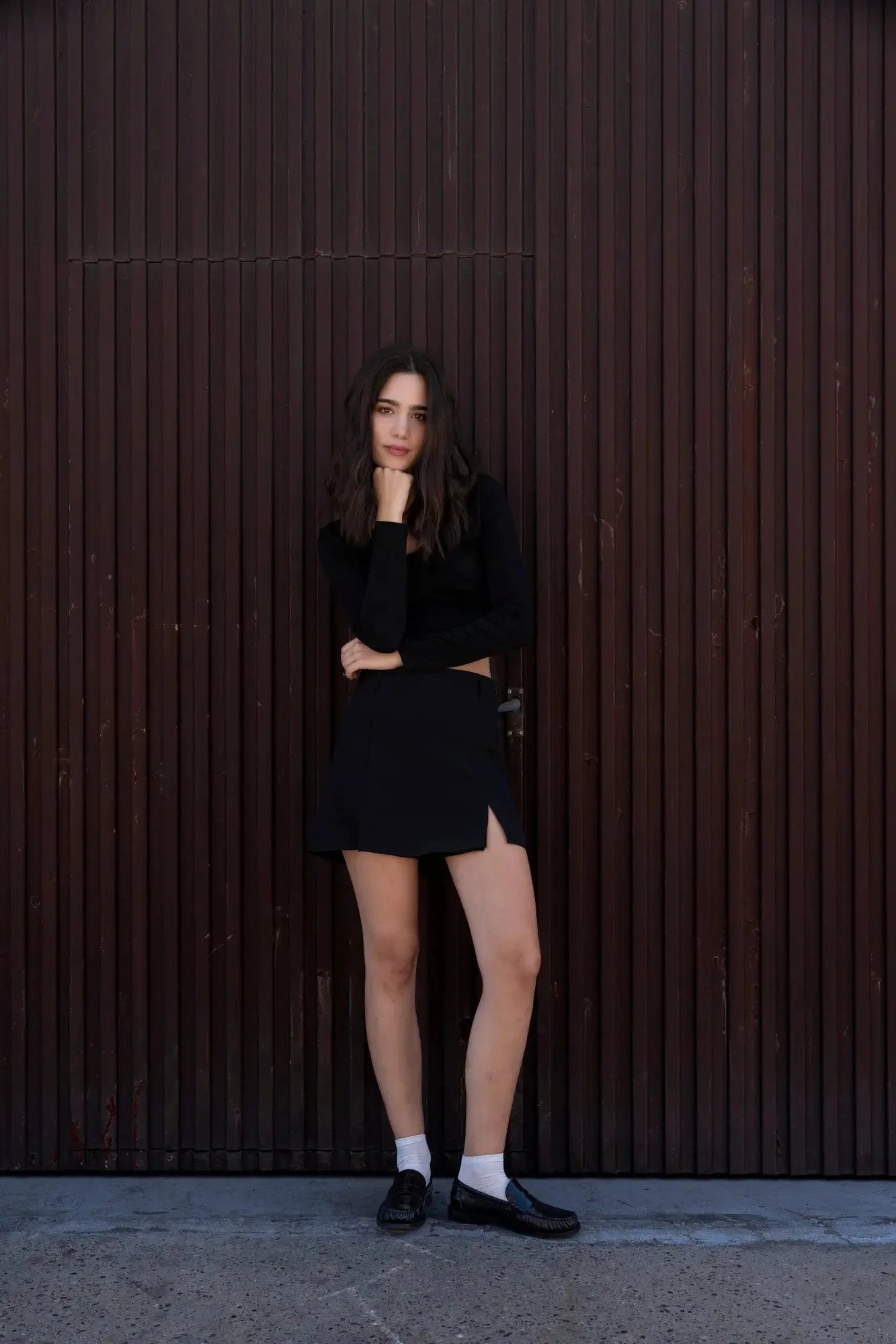 Modèle en mini jupe noir fendue, posant avec un style décontracté, mettant en valeur la jupe avec des chaussettes blanches et des chaussures noires.