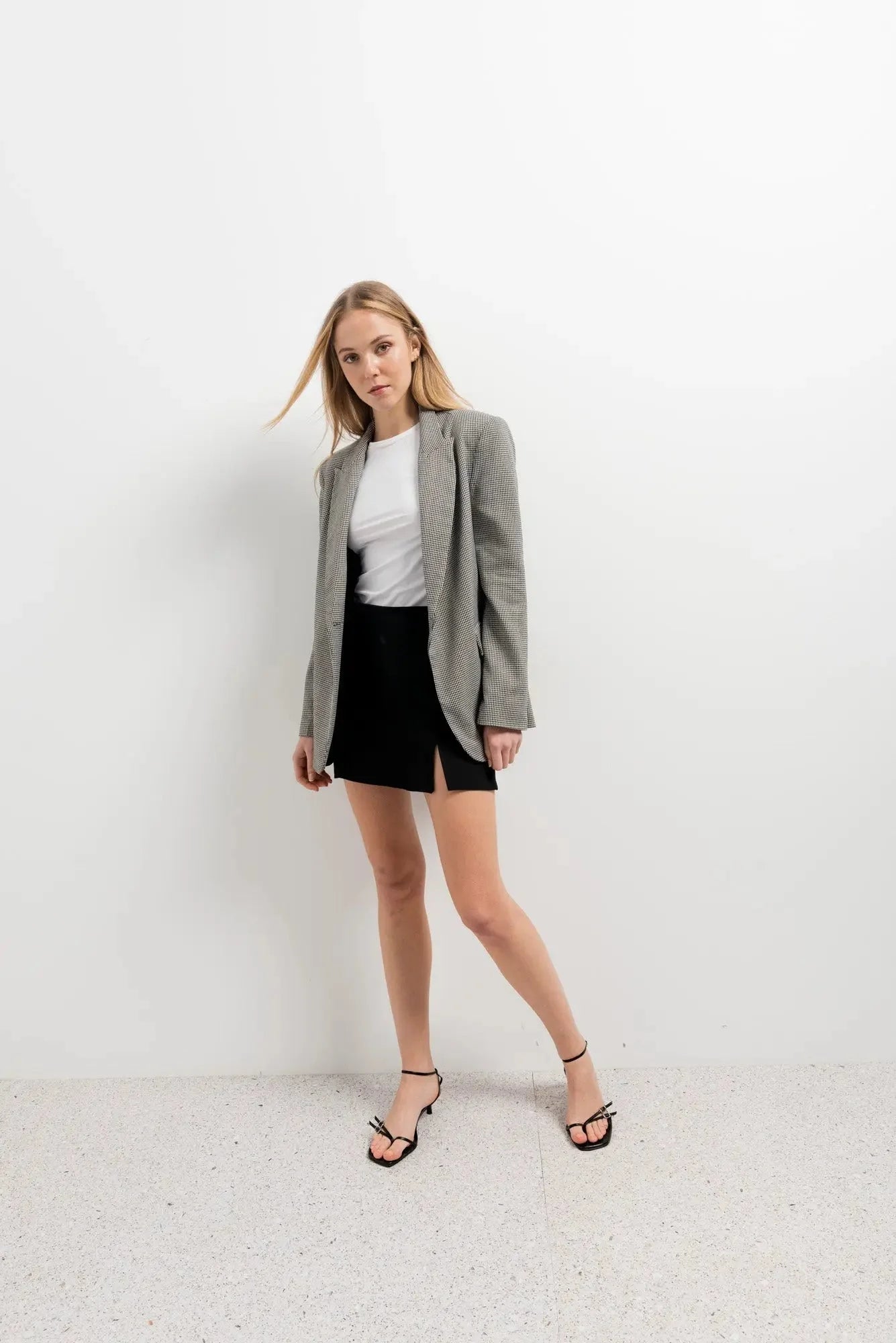 Femme stylée en mini jupe noir fendue, associée à un blazer gris et des sandales, pour un look chic et moderne.