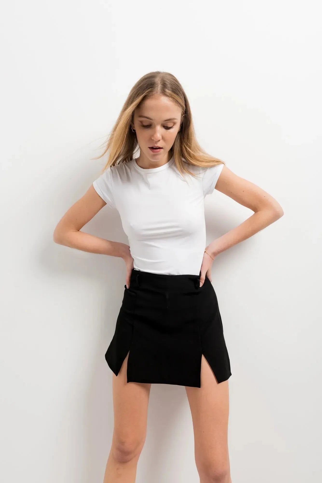 Femme en mini jupe noir fendue, posant avec un t-shirt blanc, soulignant le design élégant de la jupe.