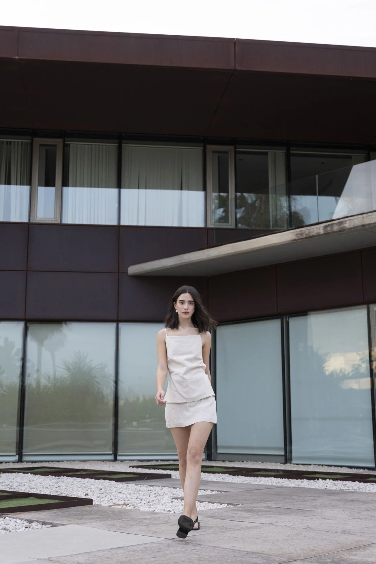 Modèle marchant dans une mini jupe short en lin naturel, 100% léger et respirant, devant un bâtiment moderne.