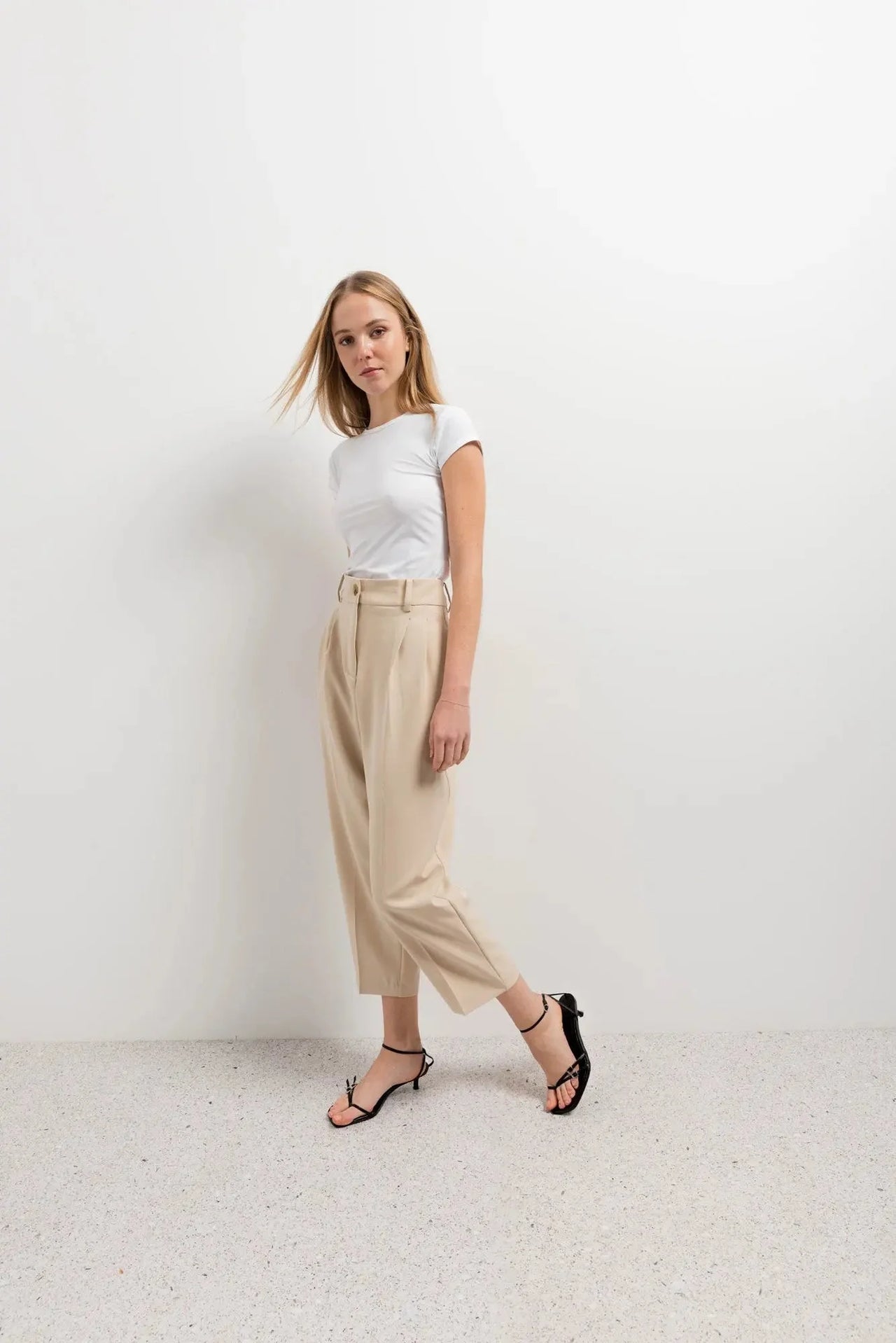 Femme stylée en t-shirt blanc et pantalon court plissé taille basse beige, illustrant un look décontracté élégant avec le pantalon court noir femme en tête.