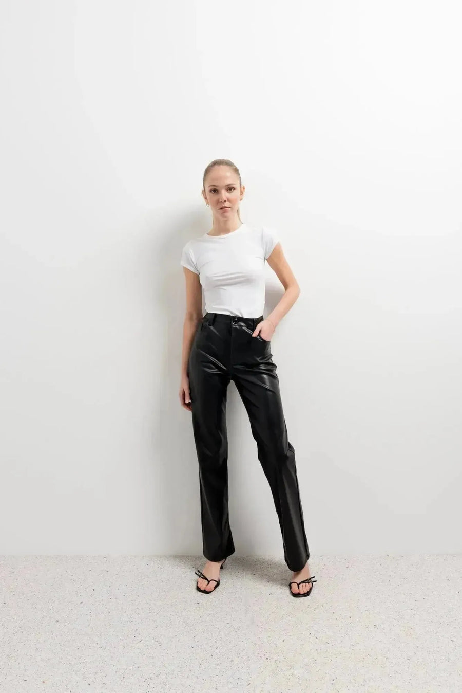 Femme posant avec assurance dans le Pantalon Effet Cuir Noir Élégant, associé à un top blanc. Idéal pour toutes celles qui recherchent un pantalon imitation cuir femme.