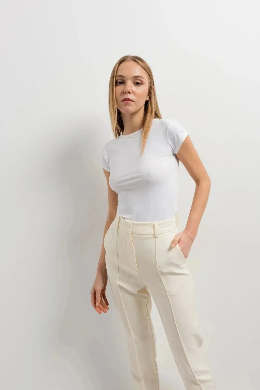 Femme portant un pantalon habillé femme écru lumineux, style chic, avec un haut blanc ajusté, posé contre un mur blanc.