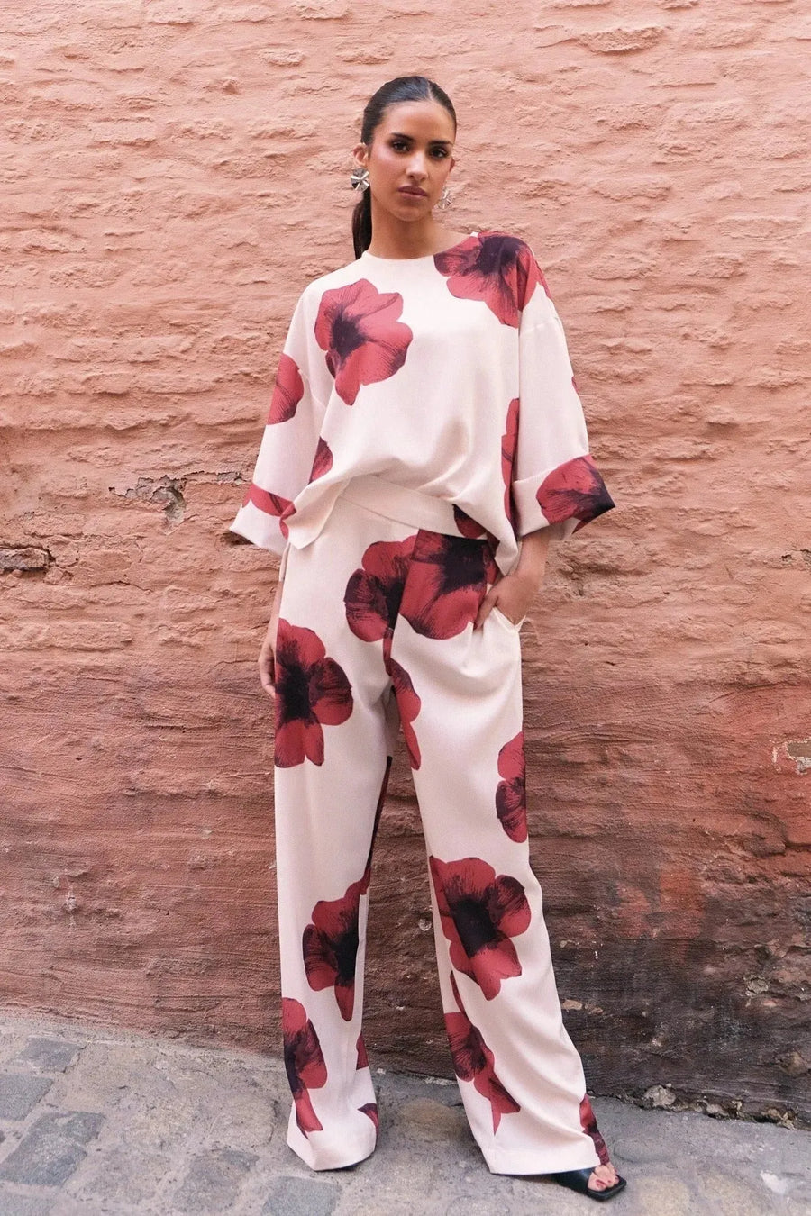 Femme souriante vêtue d'un ensemble décontracté dans un pantalon habillé fluide – Coupe droite moderne – Blanc à fleurs rouges – Matière légère, soulignant le pantalon motif femme.